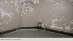 Crumbling drywall in a small room with black trim and one utilitarian chair on an exposed floor.
